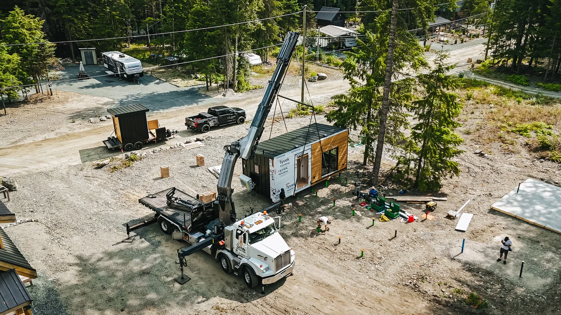 A cabin module craned from its delivery truck onto a forested site, crew setting the foundation posts